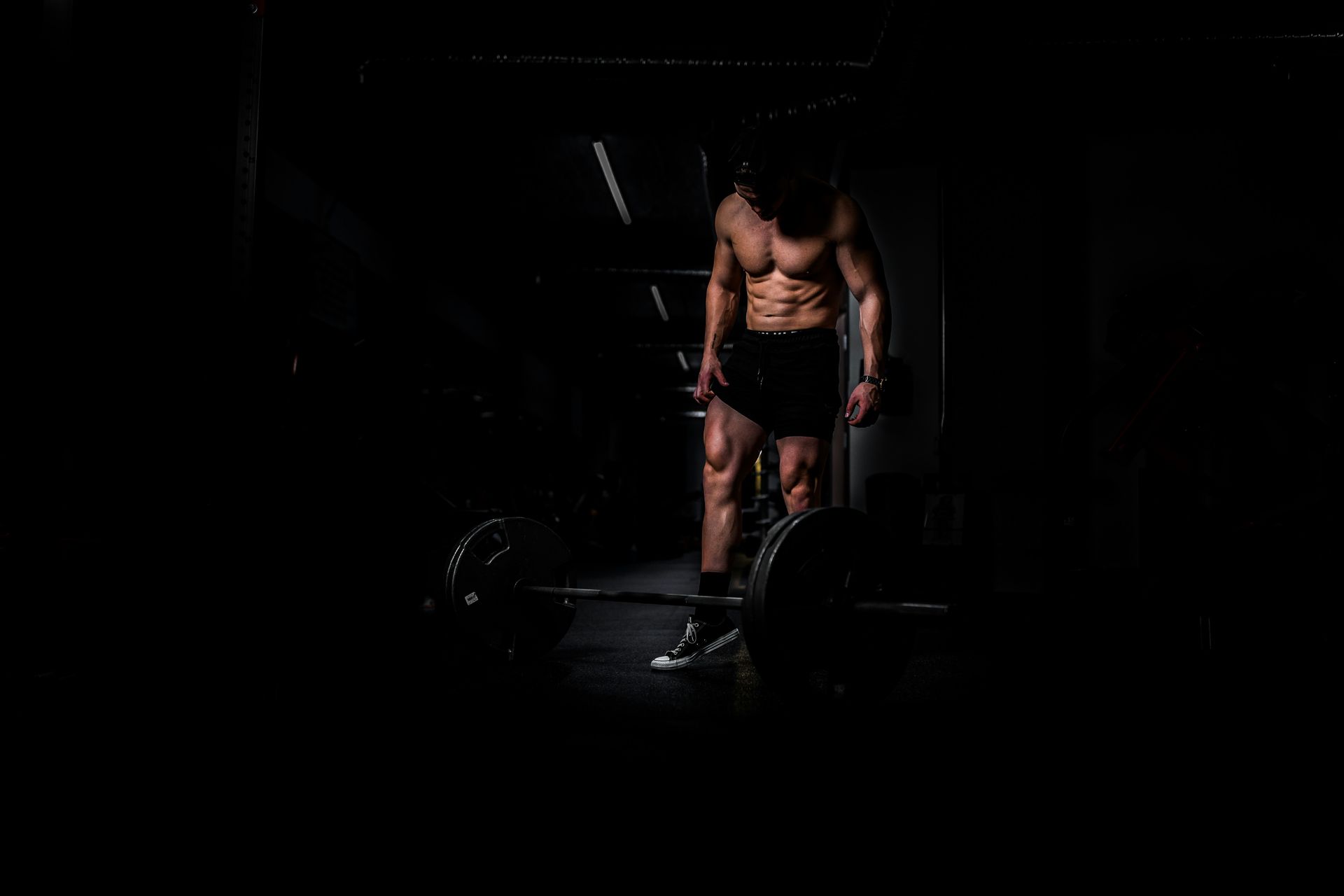 A shirtless man in black shorts standing by a barbell in a dark gym